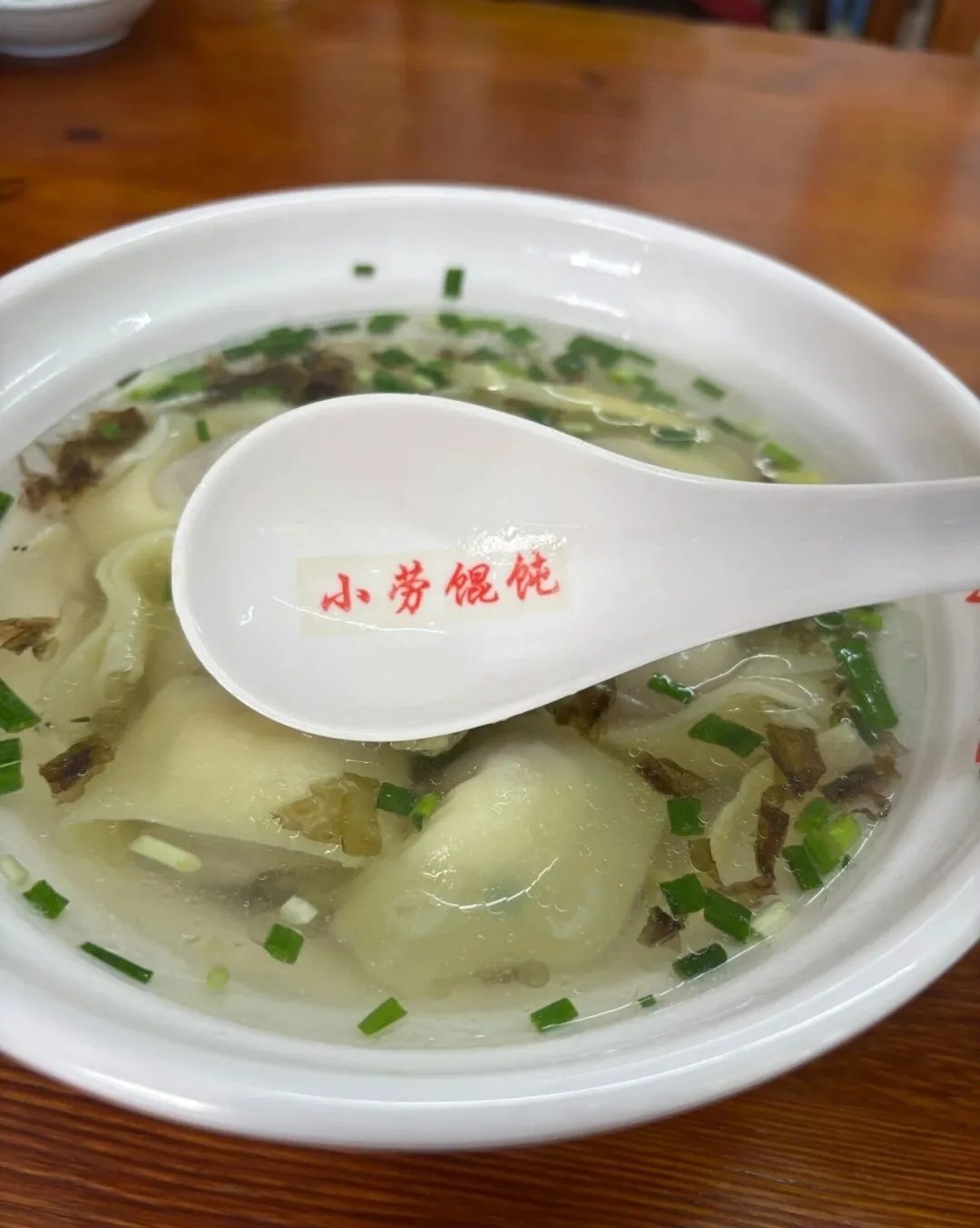 A bowl of wonton soup with greens and a Xiao Lao Hun Tun spoon