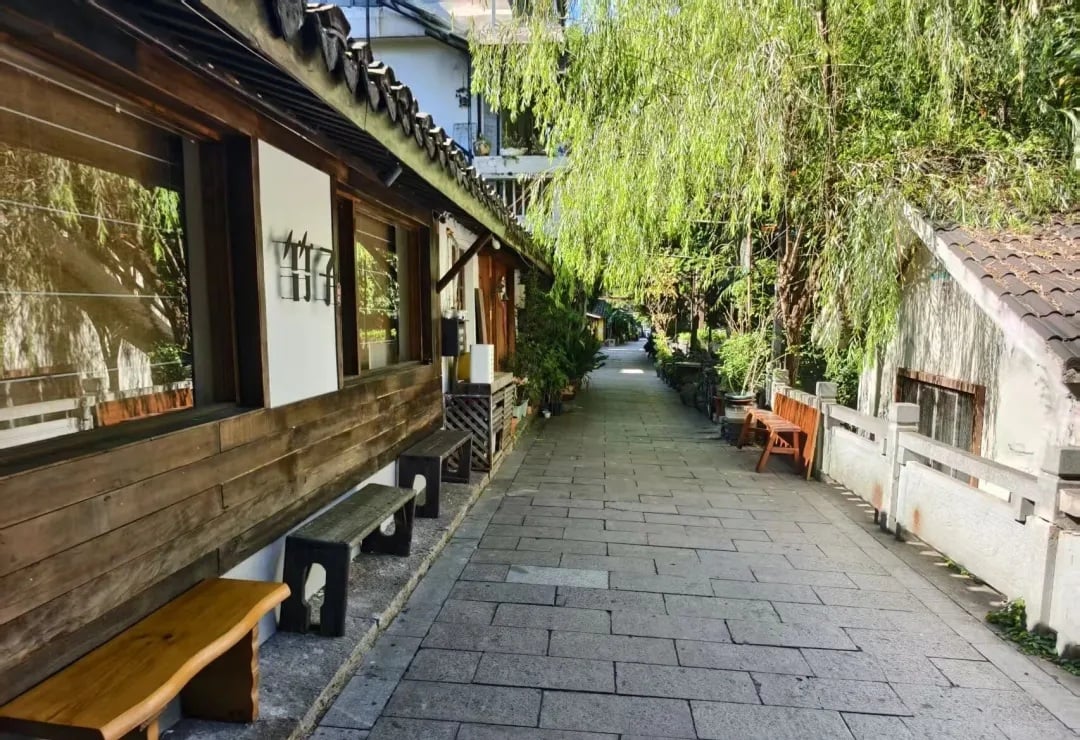 A stone-paved riverside walkway beside a wooden building and weeping willows